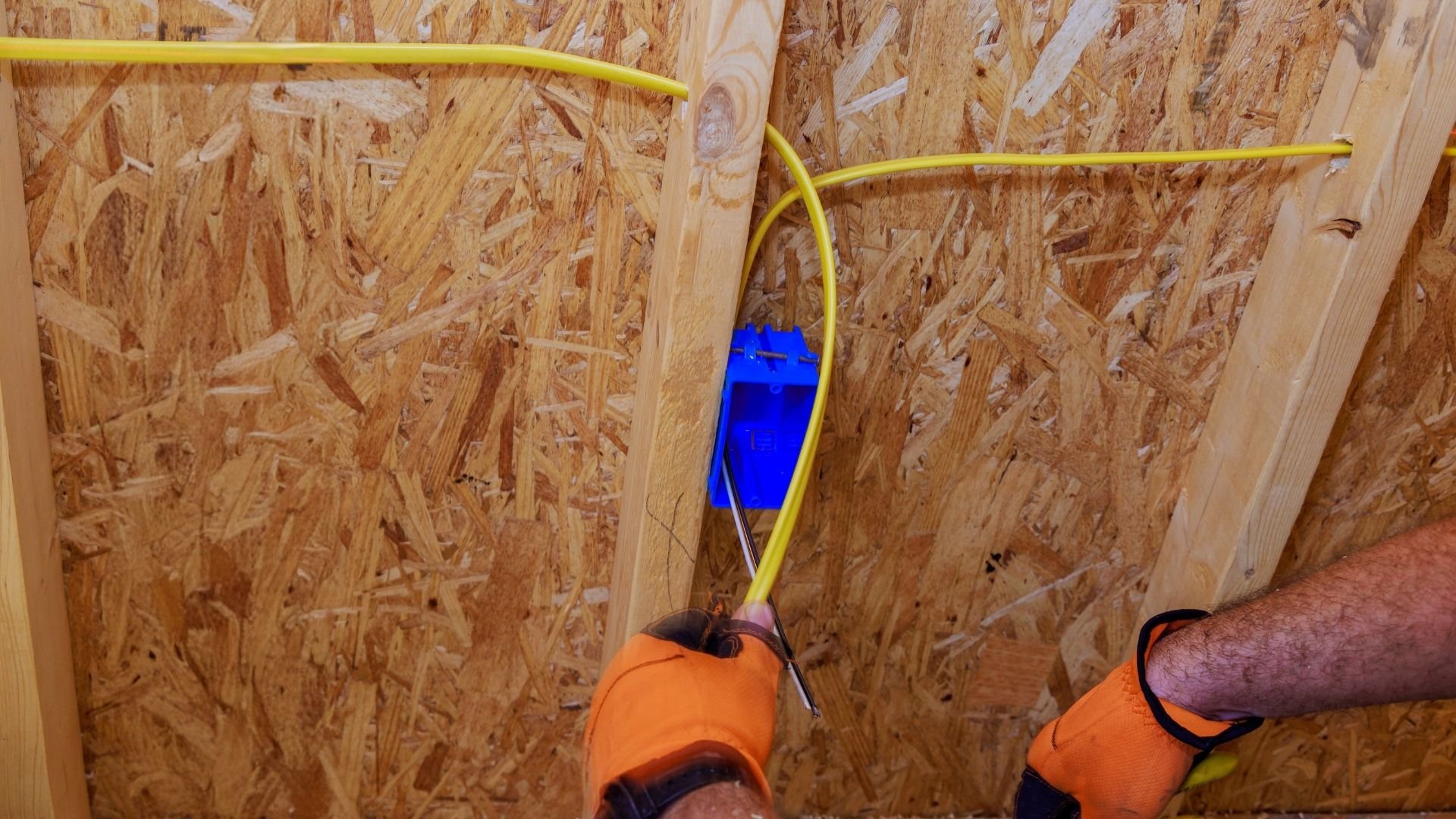 Worker installing yellow electrical conduit with blue wire against plywood wall sheathing.
