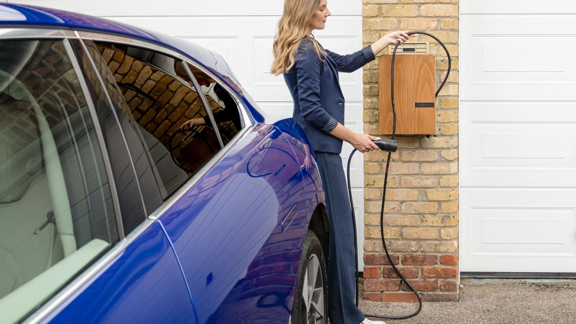 Woman charging blue electric car with home charging station on brick wall